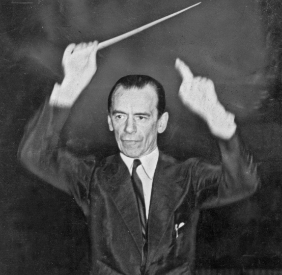 Half figure of Sir Malcolm Sargent conducting, his right arm raised holding a baton and his left arm also raised pointing his finger upwards